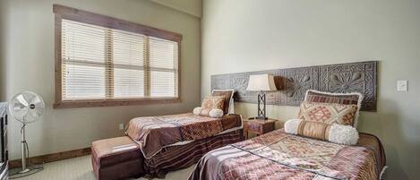 A bedroom with two beds, a large window with blinds, a standing fan, and a decorative wall panel.