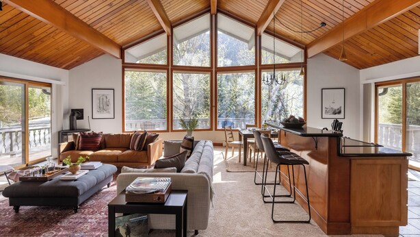 A spacious living area with a high wooden ceiling, large windows, a cozy sofa, a dining area, and a kitchen counter. - Bighorn by Avantstay Spacious Ski Cabin w/ Sprawling Porch, Views & Hot Tub! (Vail)