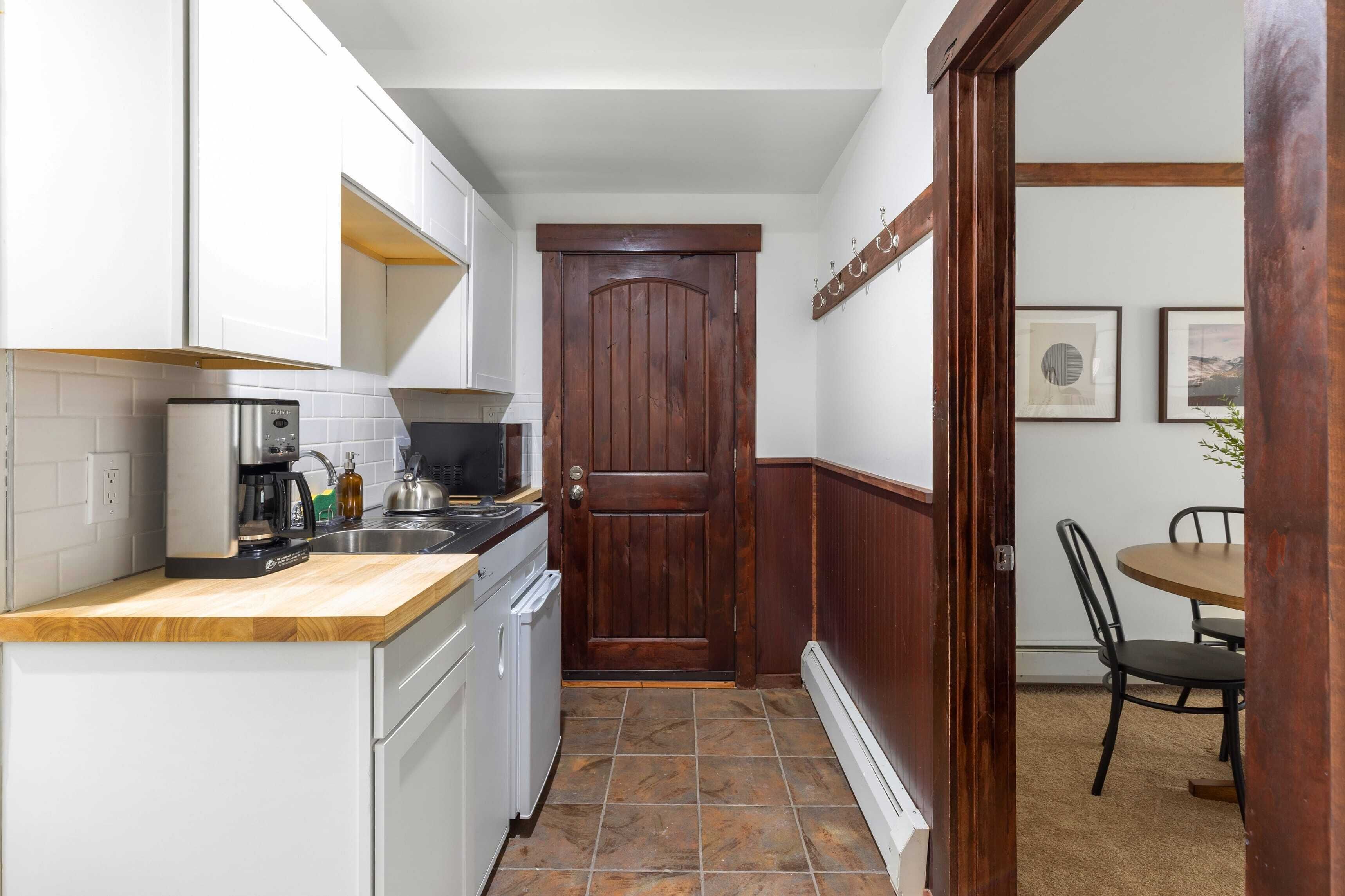 A compact kitchen with white cabinets, a wooden countertop, a built-in oven, and a door leading to a dining area with a round table and chairs.