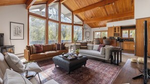 A spacious living area with large windows, a fireplace, and a kitchen in the background. - Bighorn by Avantstay Spacious Ski Cabin w/ Sprawling Porch, Views & Hot Tub! (Vail)