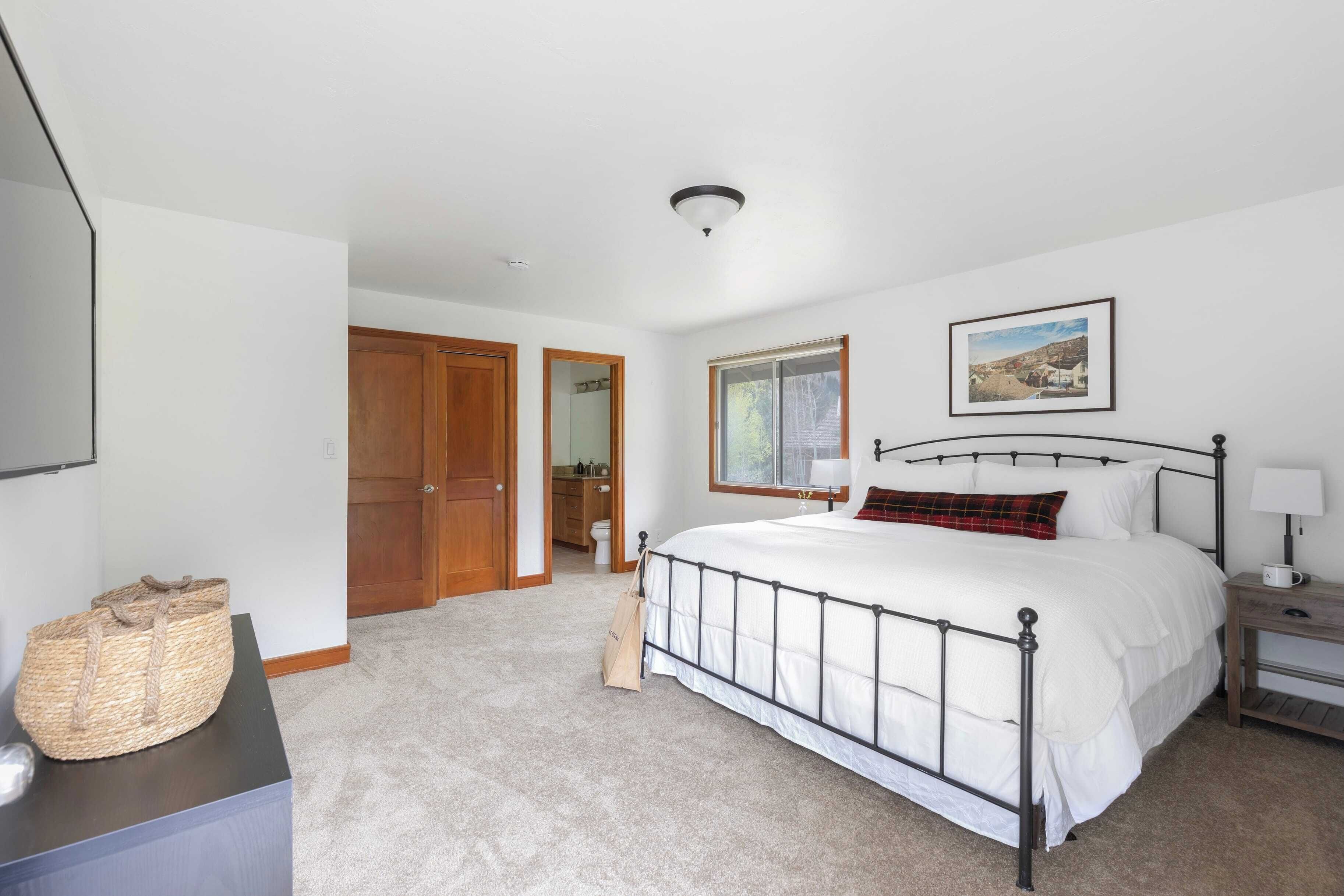 A bedroom with a bed, a nightstand, a lamp, a window, and a door leading to a bathroom.
