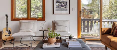 Una sala de estar con un sofá, sillones, una mesa de centro con libros y una planta, una guitarra y un cuadro enmarcado de una estación de esquí.
