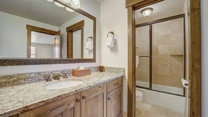 A bathroom with a marble countertop, wooden cabinets, a large mirror, and a glass-enclosed shower.