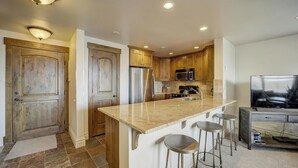 A kitchen with wooden cabinets, a stainless steel refrigerator, a microwave, a granite countertop, and a bar stool. - Resort Plaza #5061 by Avantstay Located in Park City Mountain Resort Close to the Slopes! (Park City)