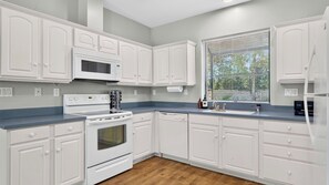Une cuisine avec des meubles blancs, une cuisinière, un four à micro-ondes et une fenêtre avec des stores.
