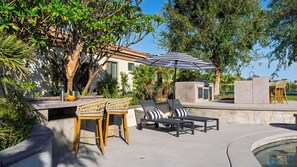 Une terrasse extérieure avec une piscine, des chaises longues, une table avec des boissons et un grand parasol.