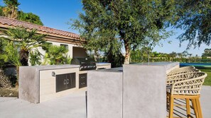 A patio area with a barbecue grill, a concrete seating area, and a pool in the background. - Villa Verbena by Avantstay Expansive Estate w/ B-ball Court, Pool/spa & Entertainer's Yard (Indio)