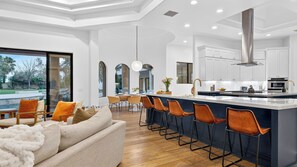A modern kitchen with white cabinets, a stainless steel range hood, and a bar area with stools. - Villa Verbena by Avantstay Expansive Estate w/ B-ball Court, Pool/spa & Entertainer's Yard (Indio)