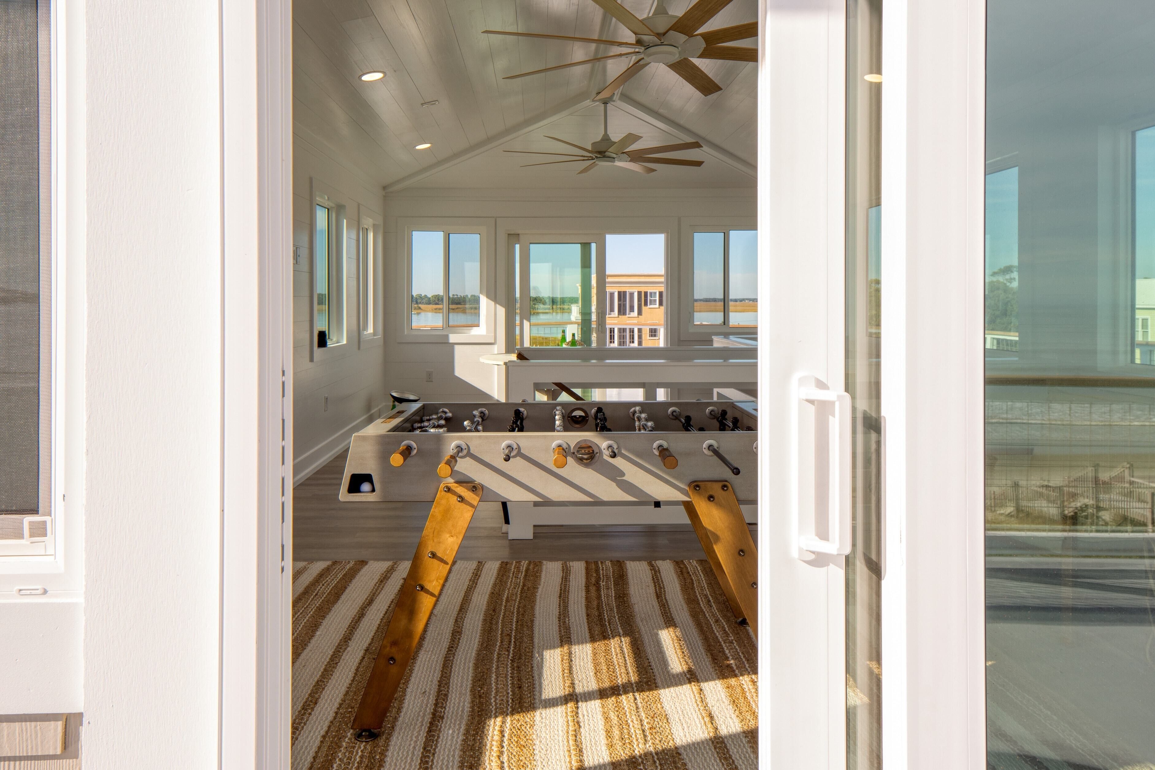 A room with a foosball table, large windows, and a ceiling fan.