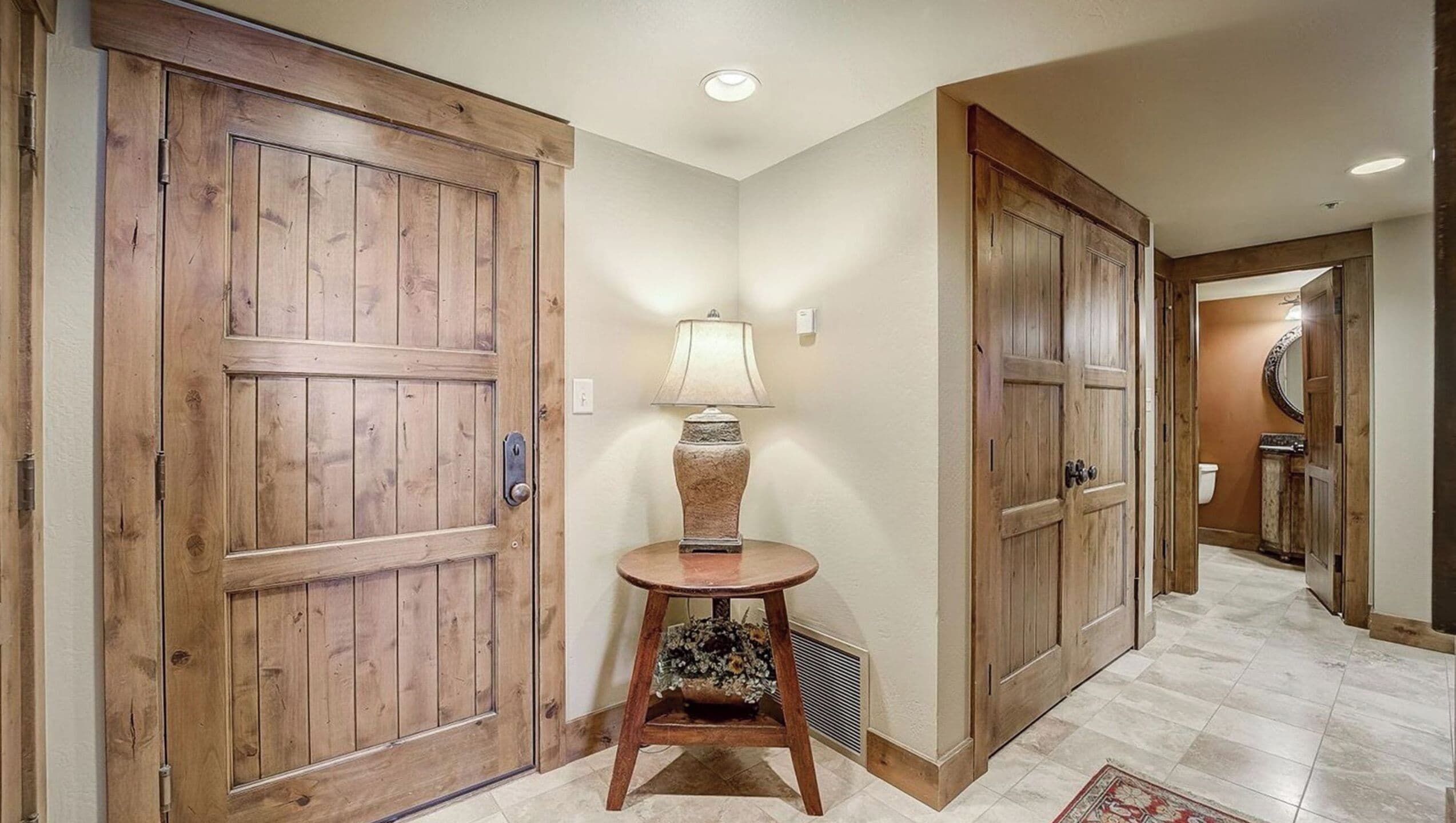 A hallway with wooden double doors, a small round table with a lamp, and a tiled floor.
