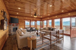 L’intérieur d’un chalet en bois est aménagé avec une cheminée, des canapés, un téléviseur et de grandes fenêtres qui offrent une vue sur l’extérieur.