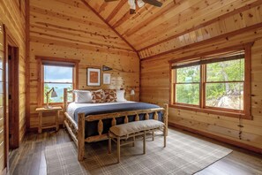 A wooden cabin bedroom with a bed, bench, bedside table, lamp, and large windows.