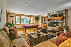 A cozy living room with a pool table, comfortable seating, a fireplace, and a large window offering a view of trees. - Telemark Luxury Retreat by Avantstay Stunning Views w/ Hot Tub, Theatre Room and Pool Table! (Park City)