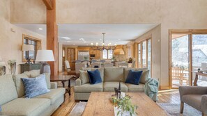 A spacious living area with a large sofa, a coffee table, and a dining table. There is a kitchen in the background with wooden cabinets and a chandelier. - Telemark Luxury Retreat by Avantstay Stunning Views w/ Hot Tub, Theatre Room and Pool Table! (Park City)