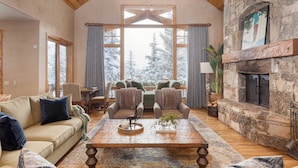 A cozy living room with a stone fireplace, wooden ceiling, and large windows with curtains.