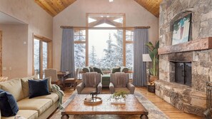 A cozy living room with a fireplace, large windows, and a stone wall. - Telemark Luxury Retreat by Avantstay Stunning Views w/ Hot Tub, Theatre Room and Pool Table! (Park City)