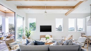A modern living room with a grey sofa, wooden chairs, a flat-screen TV, and large windows offering a view of the outdoors. - Rendezvous by Avantstay Modern Haven w/ Game Room, Roof Top, Pool & Close to Beach! (Miramar Beach)