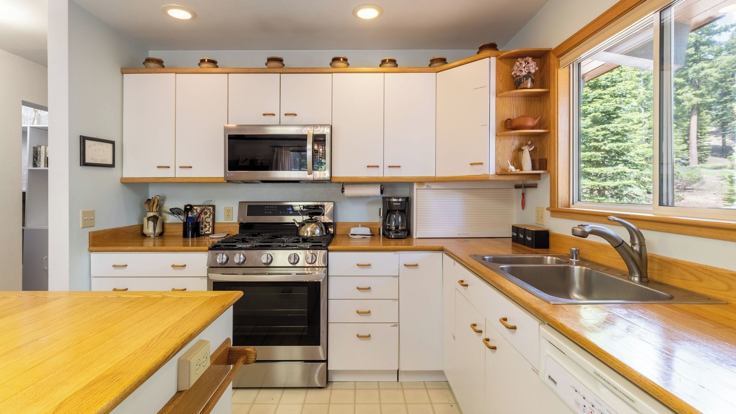 Une cuisine moderne avec des meubles blancs, des appareils en acier inoxydable, un plan de travail en bois et une fenêtre donnant sur un paysage verdoyant.