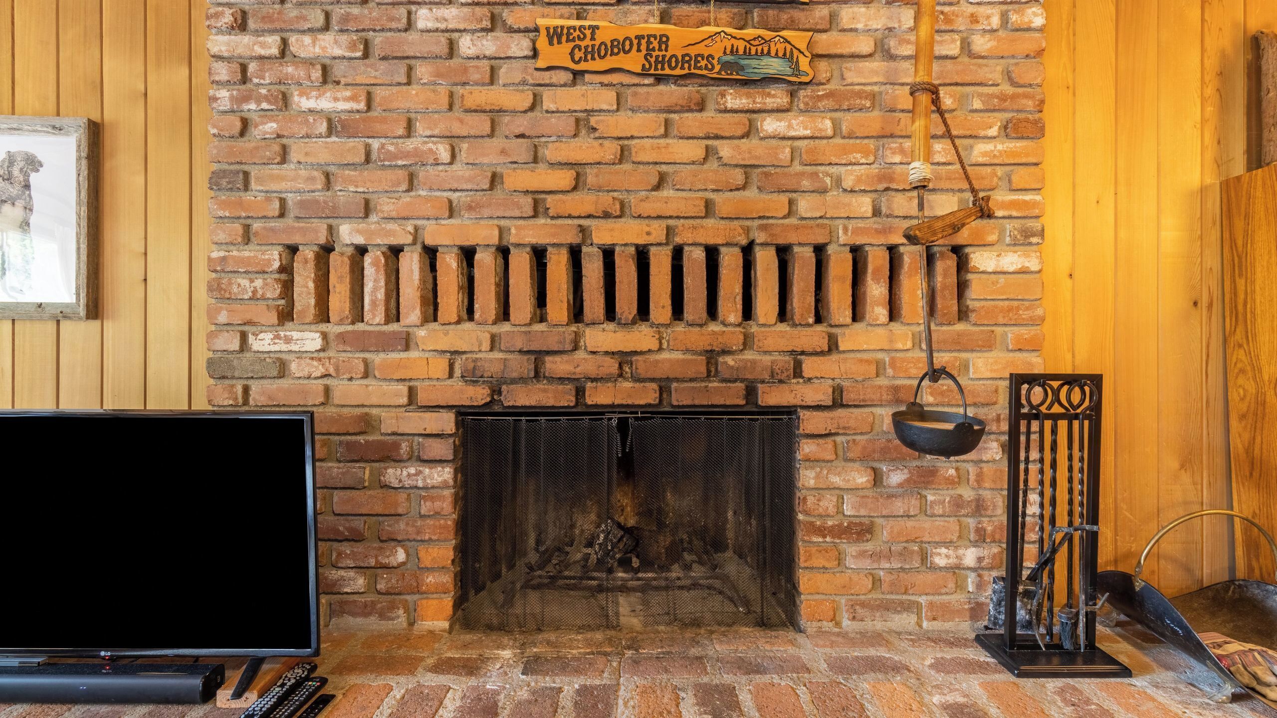 A brick fireplace with a sign above it, a TV, and a metal stand with a pot on the floor.