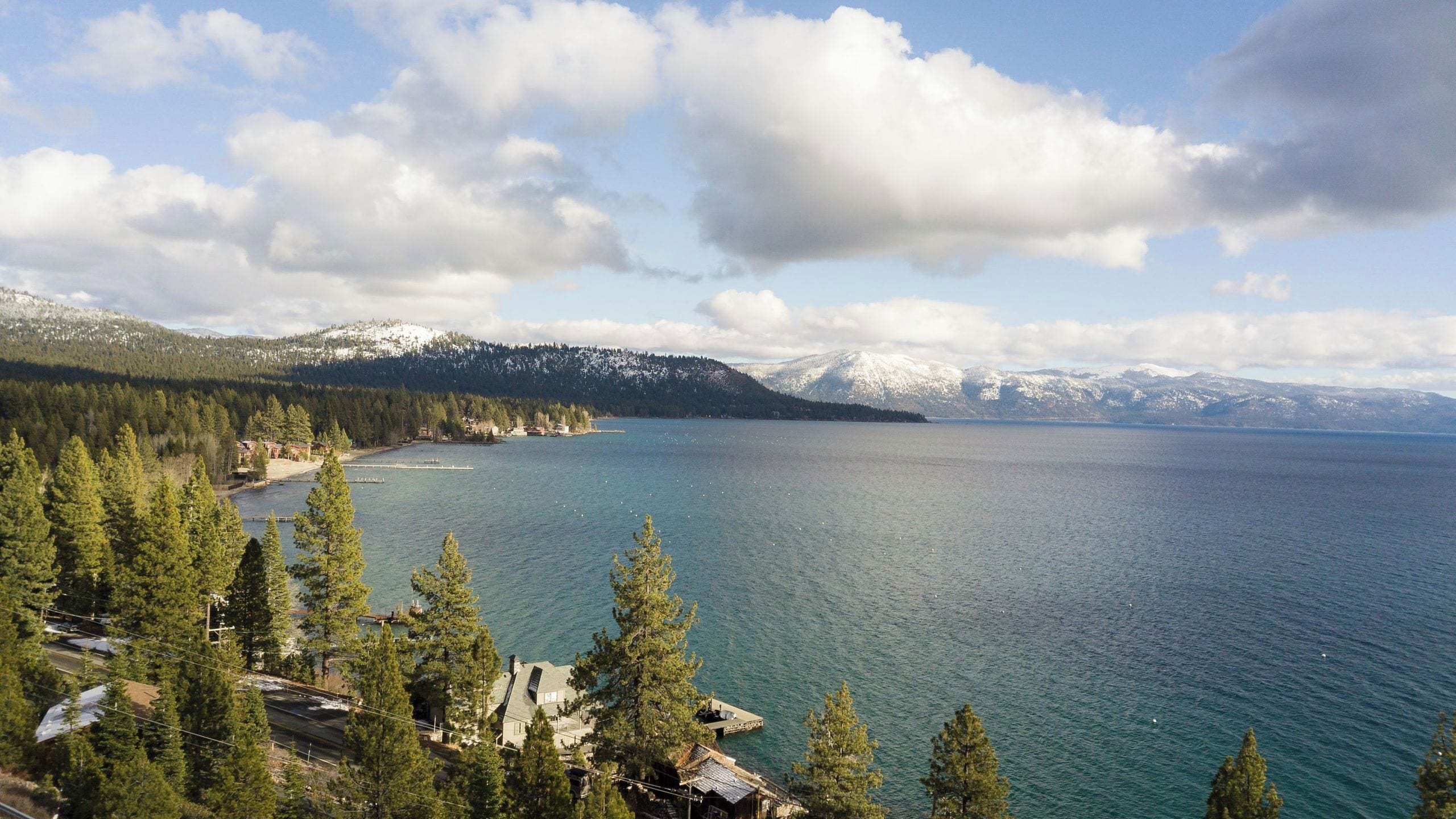 A lake with mountains in the background, surrounded by trees and a few buildings.
