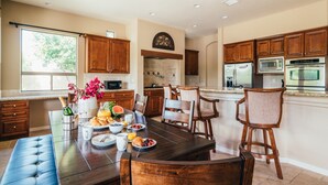 A modern kitchen with wooden cabinets, a dining table set for breakfast, and a large window providing natural light. - Desert Grove by Avantstay Oasis Retreat w/ Pool & BBQ Grill! (Indio)