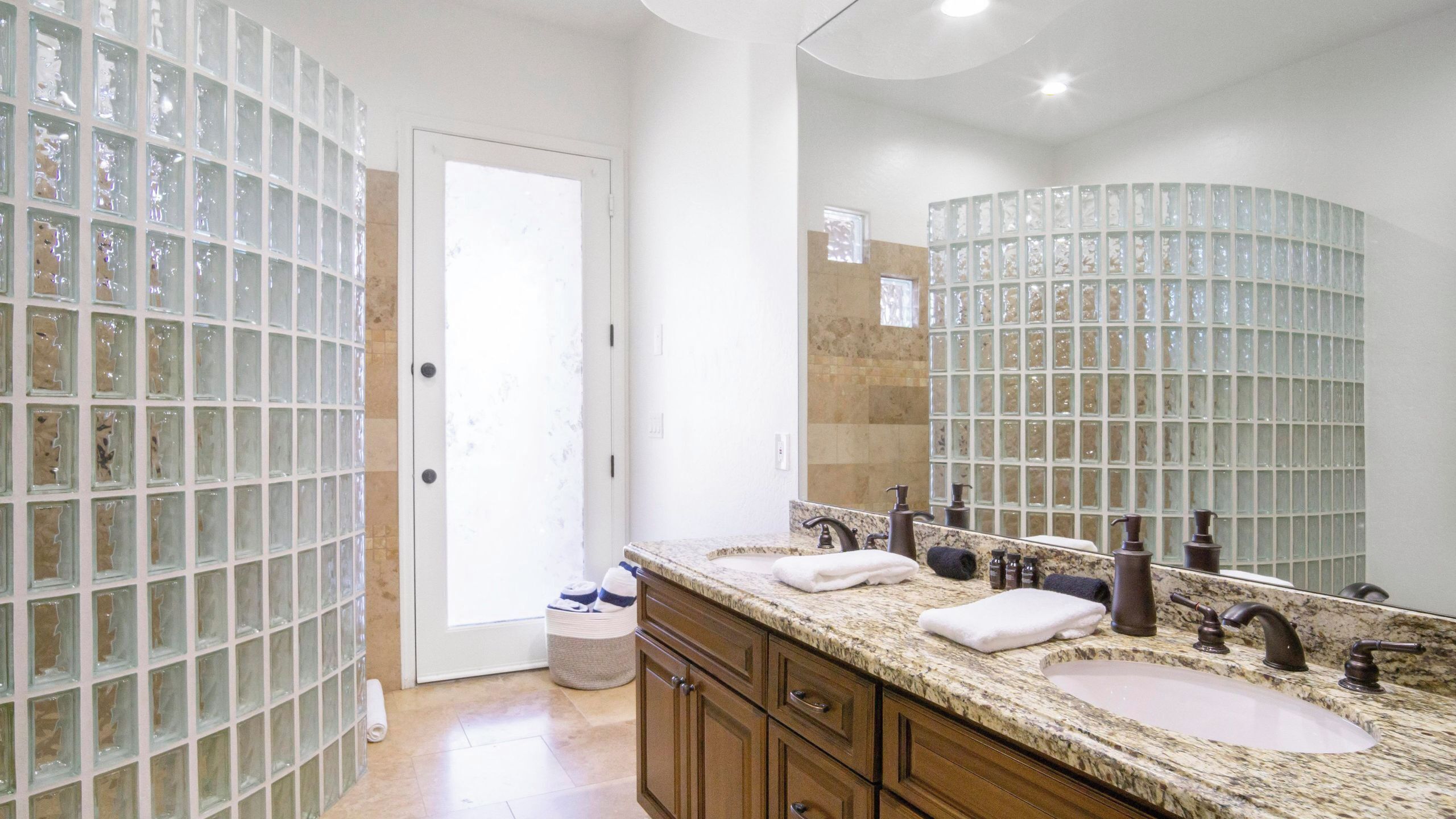 A bathroom with a double sink, two faucets, a mirror, and a glass block wall.