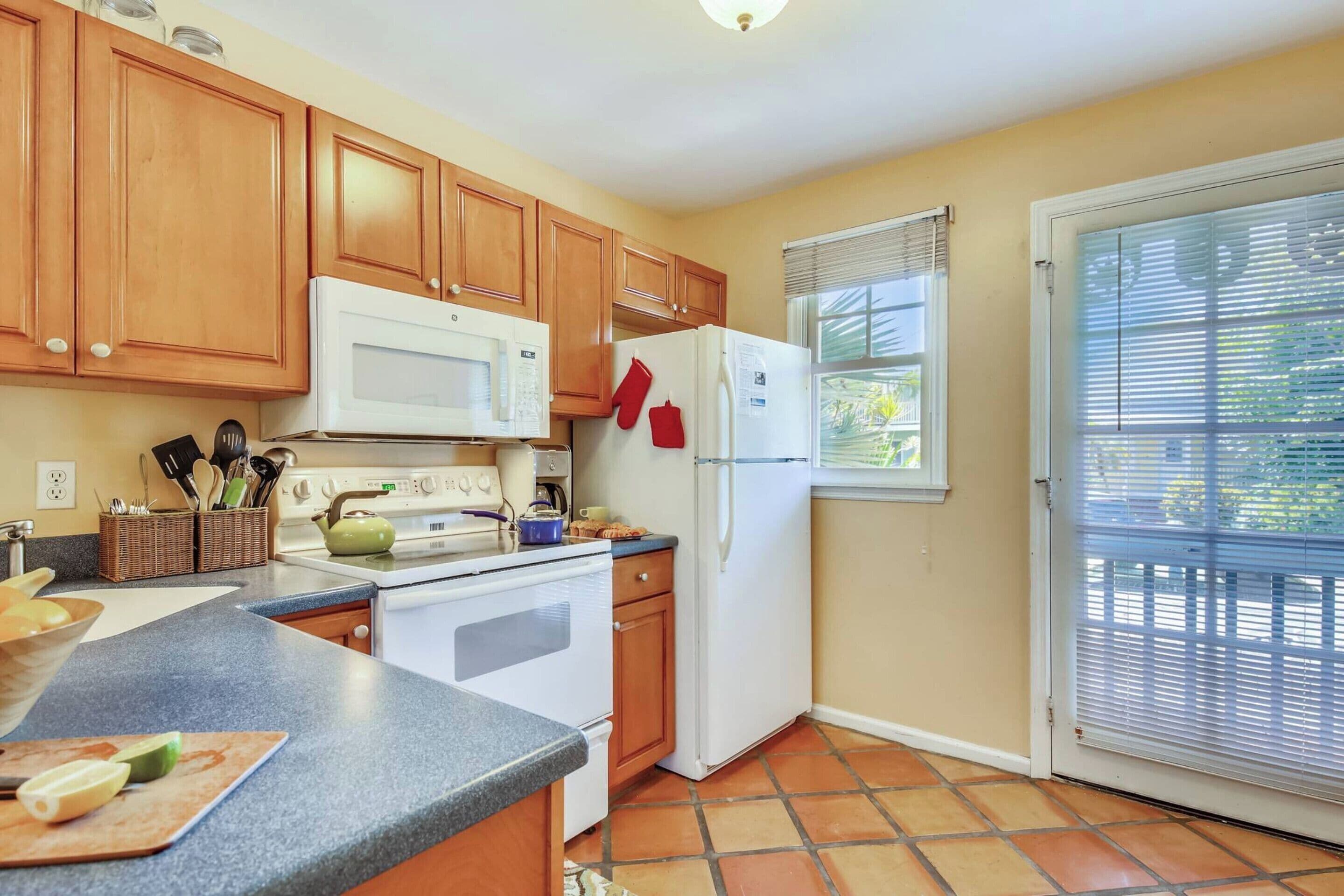 A kitchen with wooden cabinets, a white microwave, a stove, a refrigerator, and a window with blinds.