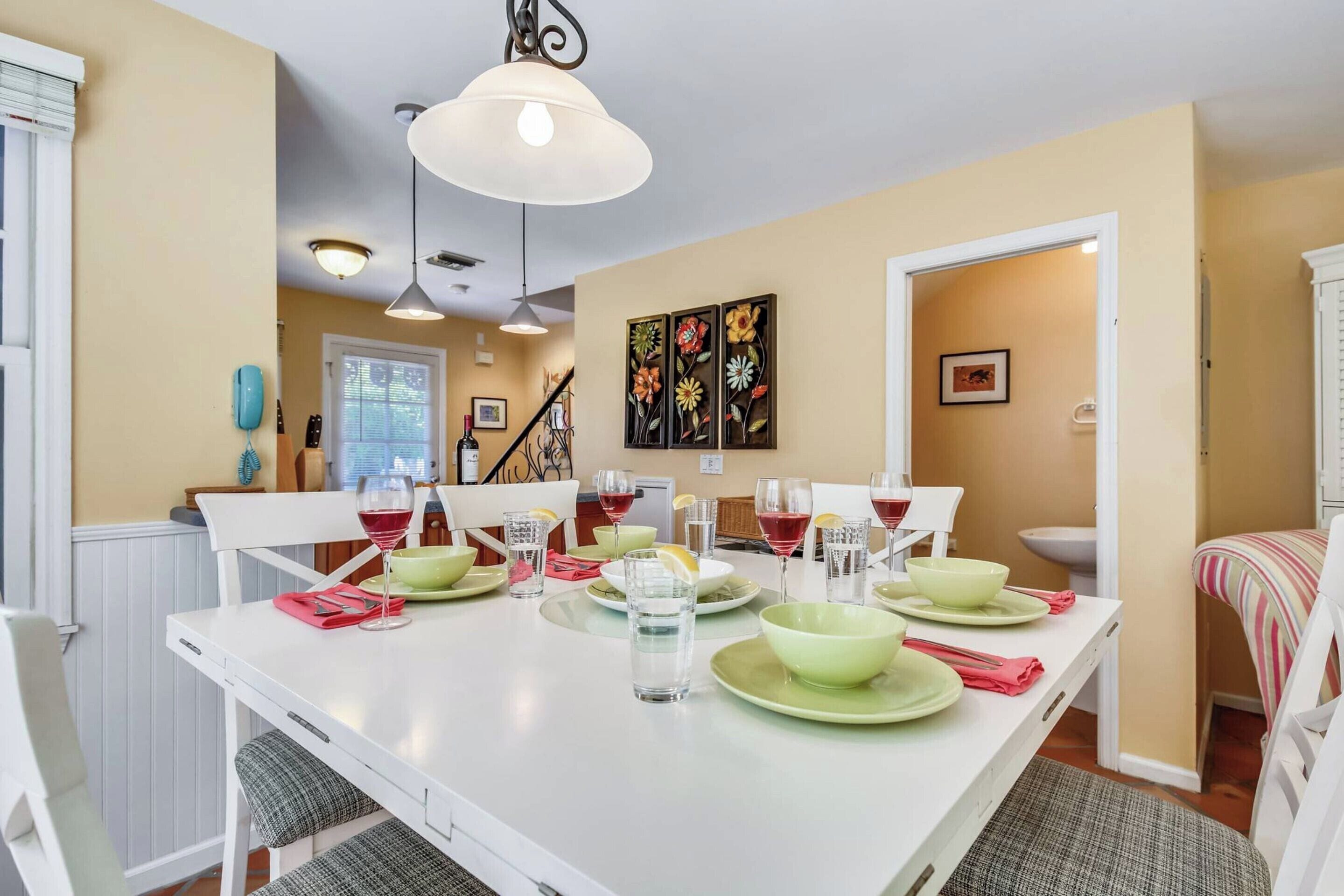 A dining area with a white table set for four, surrounded by chairs. There is a ceiling light fixture above the table.