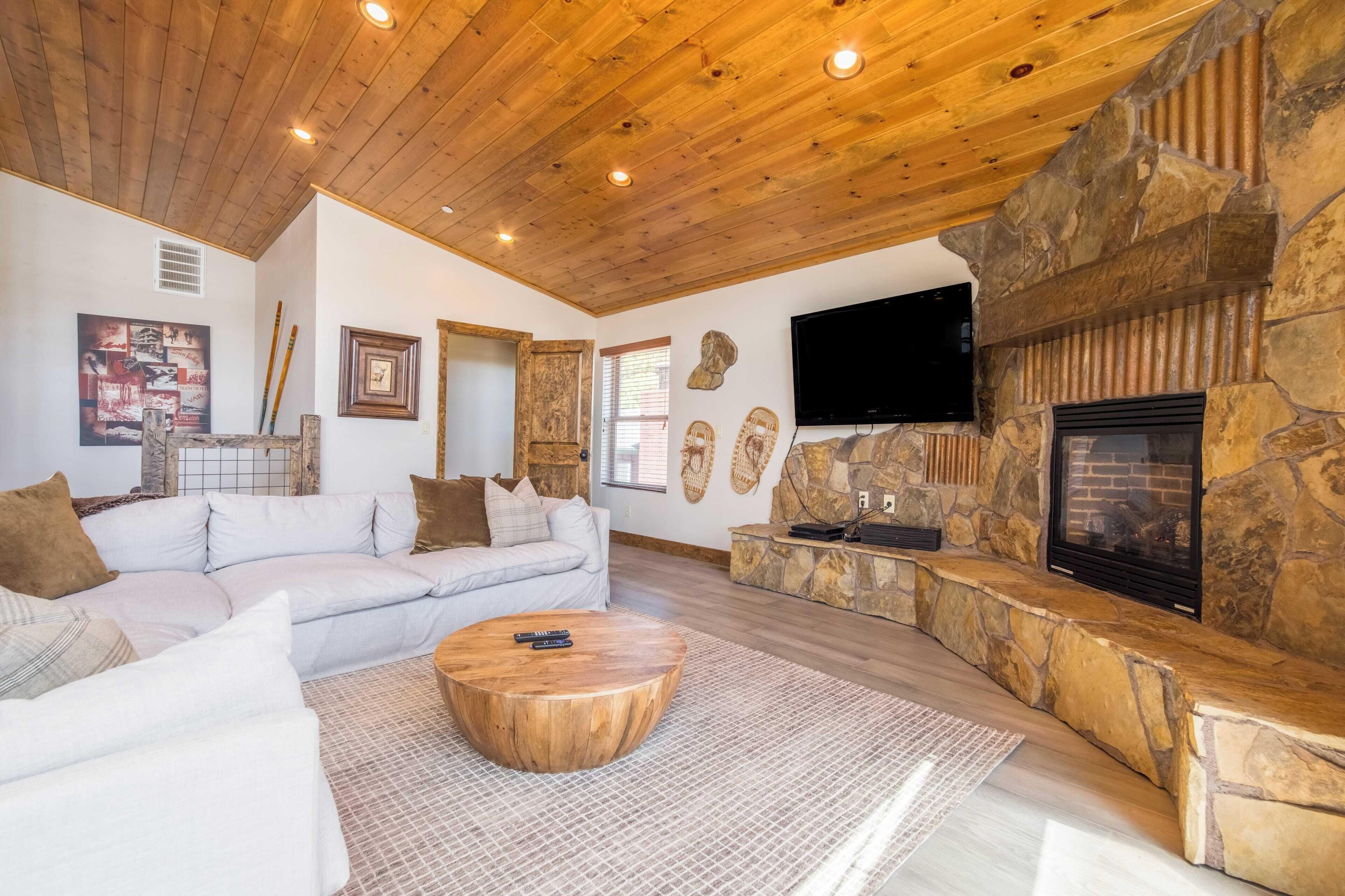A spacious living room with a stone fireplace, a large flat-screen TV, and a wooden ceiling.