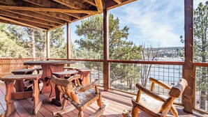 A wooden deck with rustic furniture, including rocking chairs and picnic tables, overlooking a scenic landscape with trees and a body of water. - Lakefront Lodge by Avantstay Direct Access to Big Bear Lake Hot Tub + Views (Big Bear Lake)