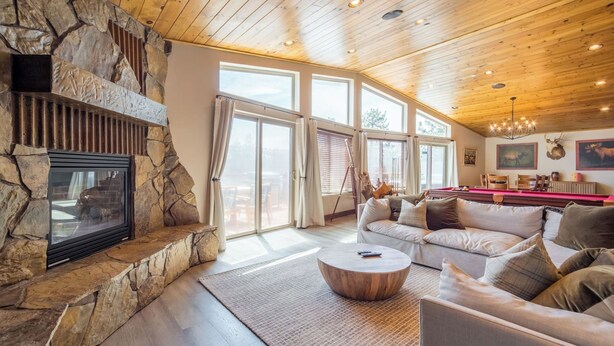 A spacious living room with a stone fireplace, large windows, a wooden ceiling, and a cozy seating area. - Lakefront Lodge by Avantstay Direct Access to Big Bear Lake Hot Tub + Views (Big Bear Lake)