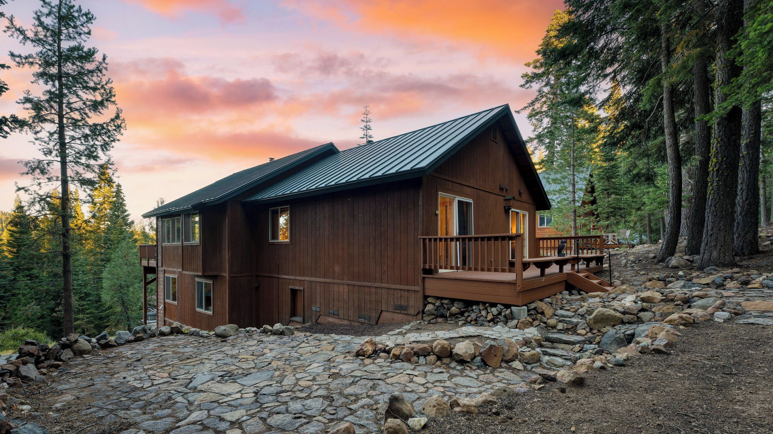 A wooden cabin with a metal roof, a deck, and a stone-paved entrance surrounded by trees.