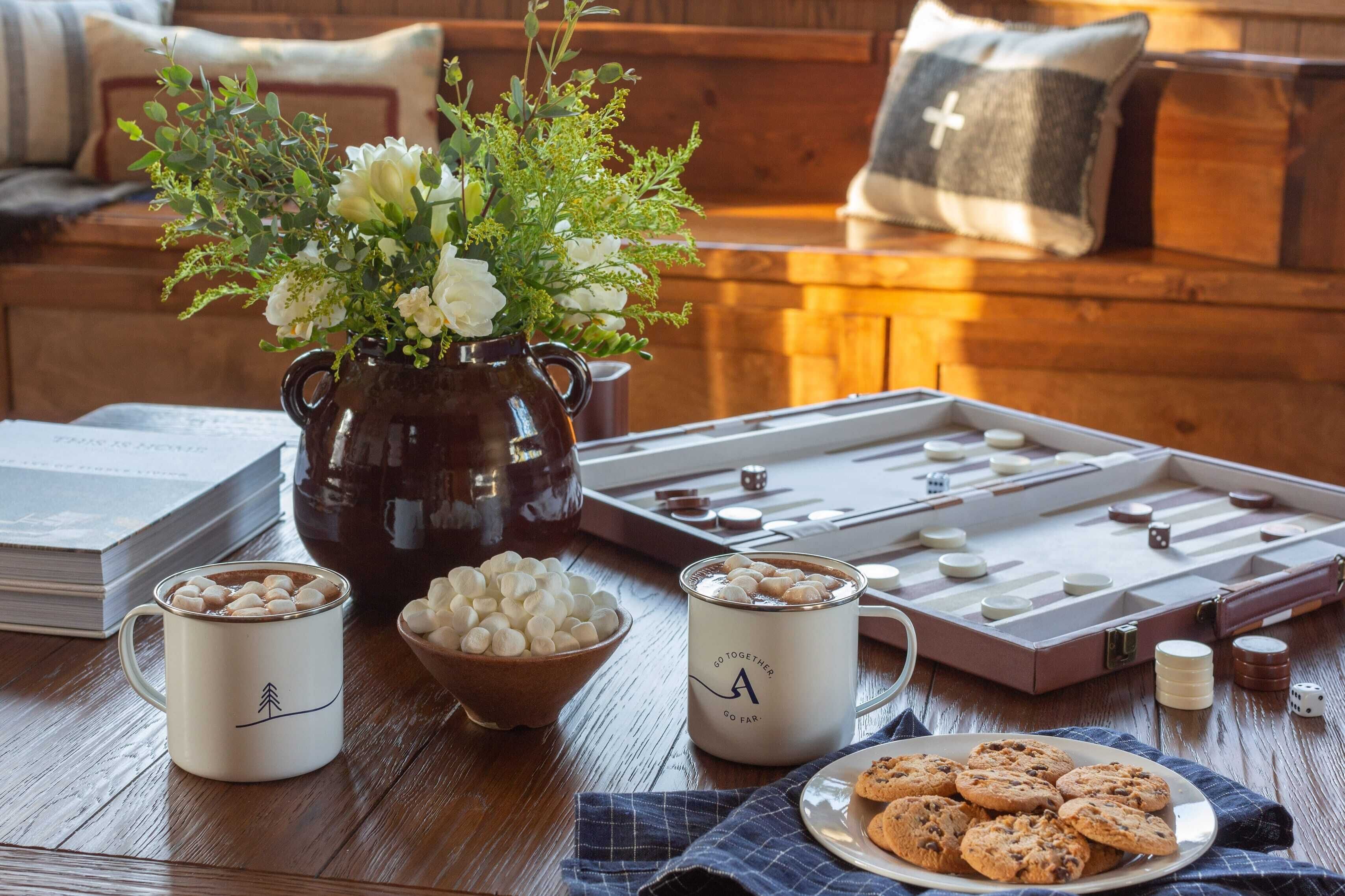Un ambiente acogedor con una mesa de madera, un tablero de ajedrez, una taza con malvaviscos, un cuenco de galletas y un jarrón con flores.