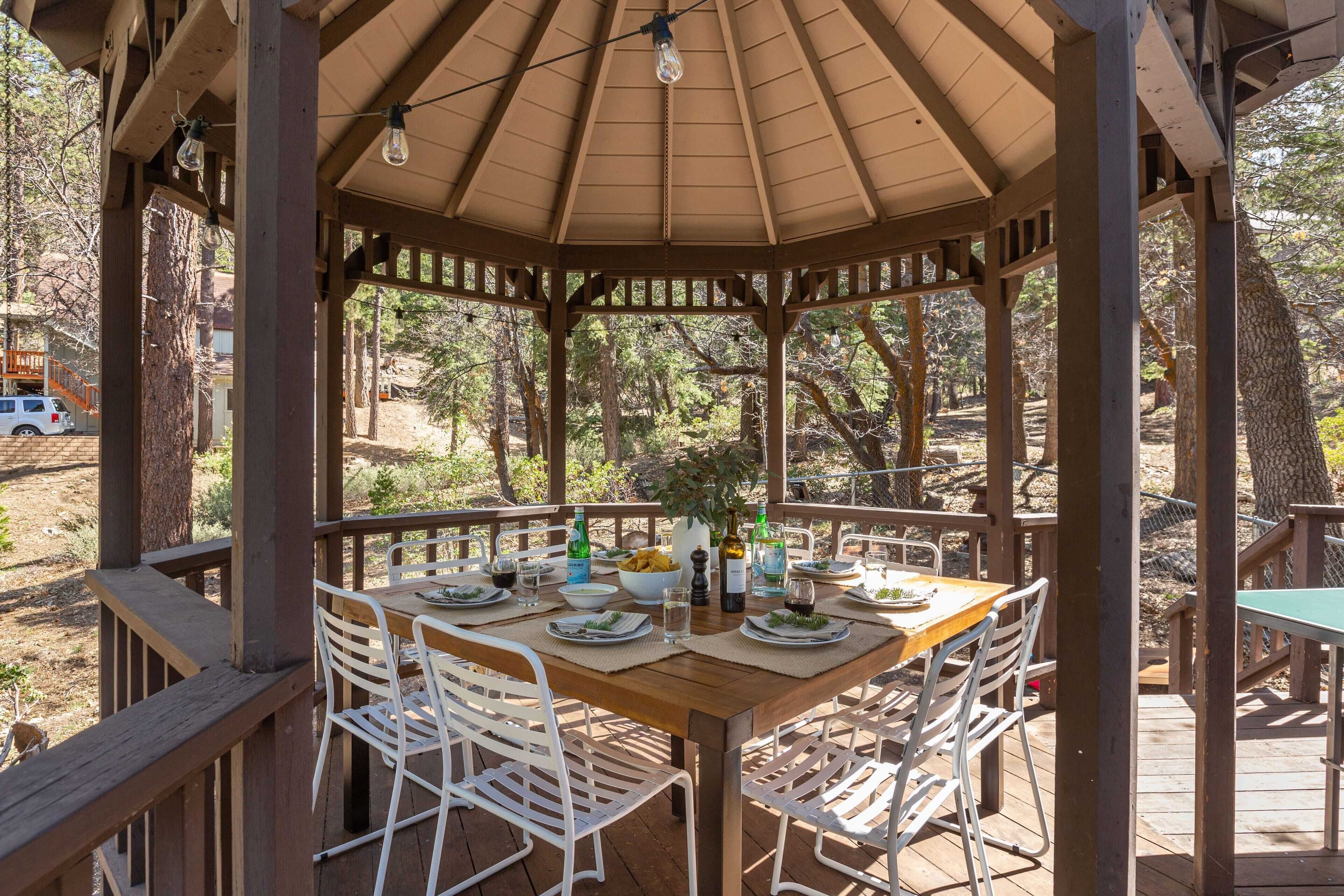A wooden gazebo with a table set for dining, surrounded by white chairs, and a view of trees and a building in the background.