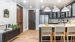 A modern kitchen with dark cabinets, a central island with chairs, pendant lighting, and a refrigerator. - Kokopelli by Avantstay Close to the Ski Slopes in This Majestic Home in Park City (Park City)