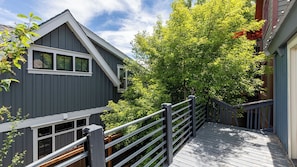 A wooden deck leading to a house with a grey exterior and a large tree in the background.