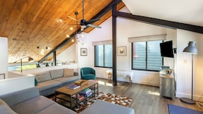 A loft-style living room with a sloped ceiling, a sofa, a coffee table, a chair, a TV, and a large window.