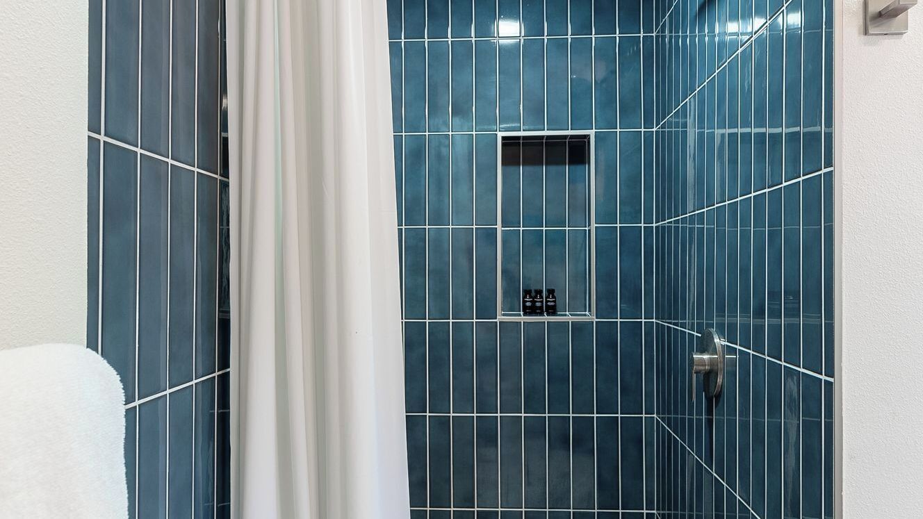 A bathroom with blue tiles, a shower area, and a small shelf with toiletries.