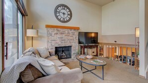 A living room with a stone fireplace, a large clock, a sofa, and a television. - Slope-side Townhome Steps to Main St, Deck View of Old Town Quittin Time Townhome by Avantstay (Park City)