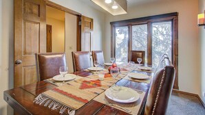 A dining room with a wooden table set for six, surrounded by leather chairs, with a view of trees through large windows. - Slope-side Townhome Steps to Main St, Deck View of Old Town Quittin Time Townhome by Avantstay (Park City)