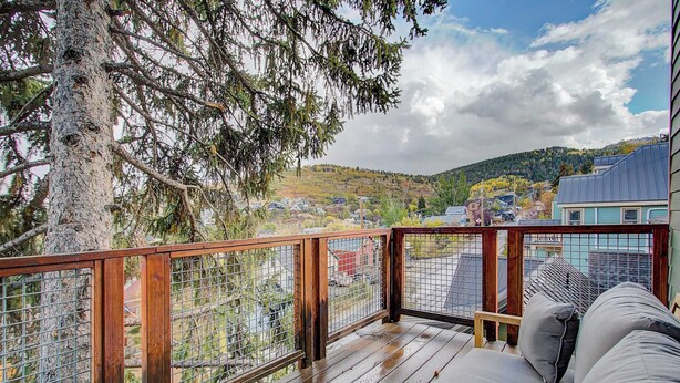 A wooden deck with a metal railing, a sofa, and a view of a town with houses and trees. - Slope-side Townhome Steps to Main St, Deck View of Old Town Quittin Time Townhome by Avantstay (Park City)