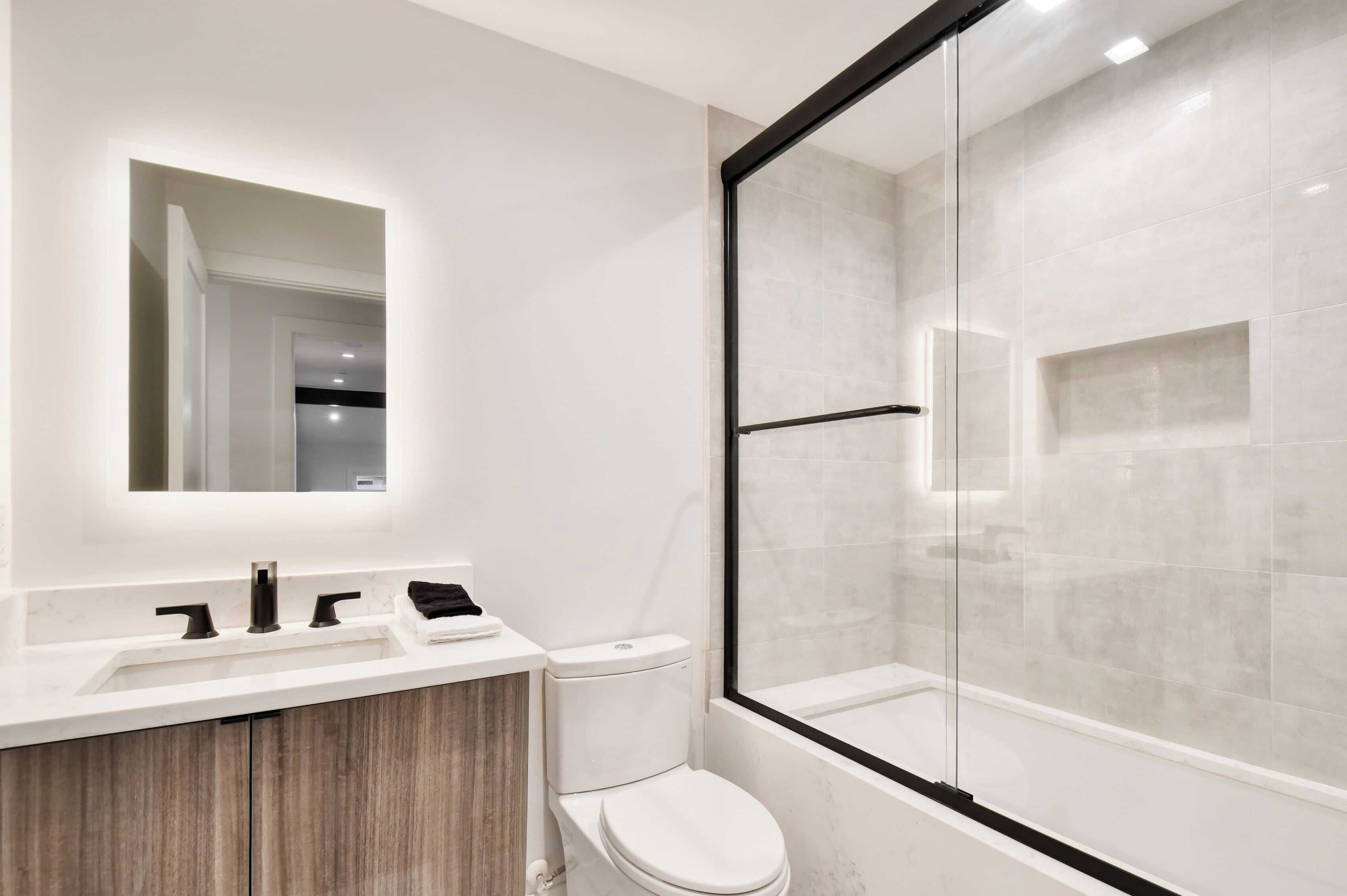 Un baño moderno con ducha de vidrio, un lavamanos blanco con tocador de madera y un inodoro.