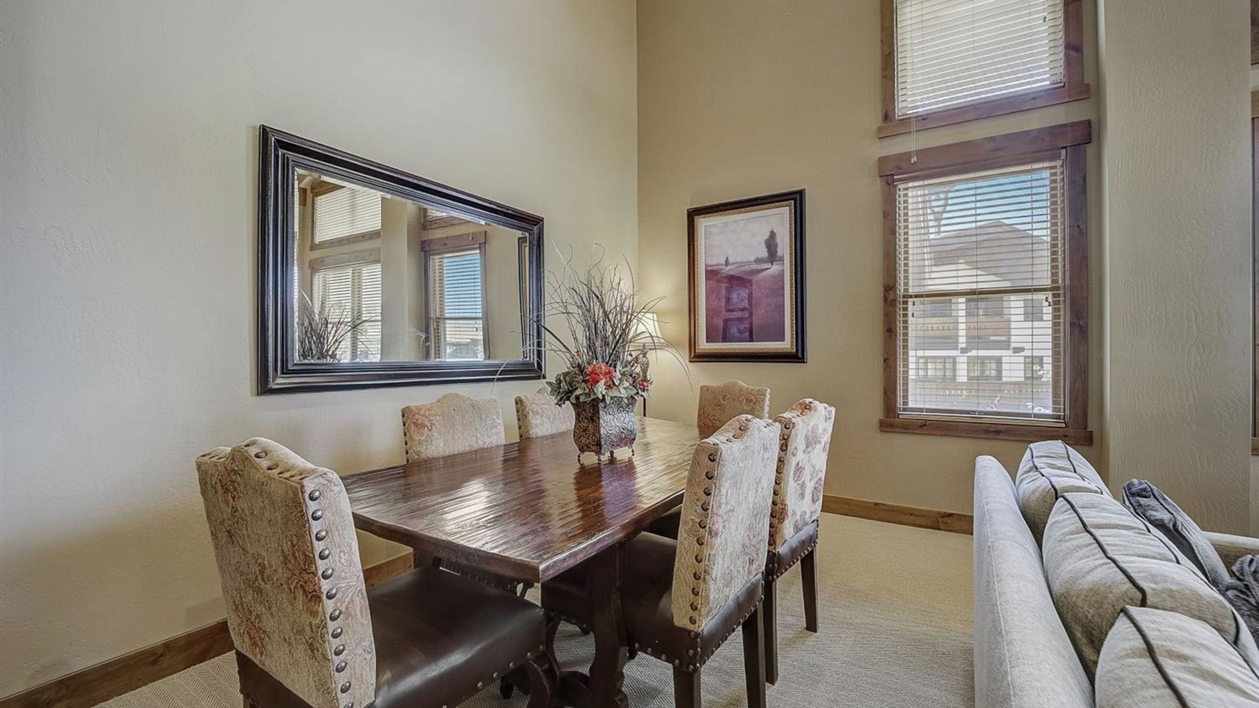 A dining area with a wooden table, six upholstered chairs, a vase with flowers, a mirror, and a framed picture on the wall.