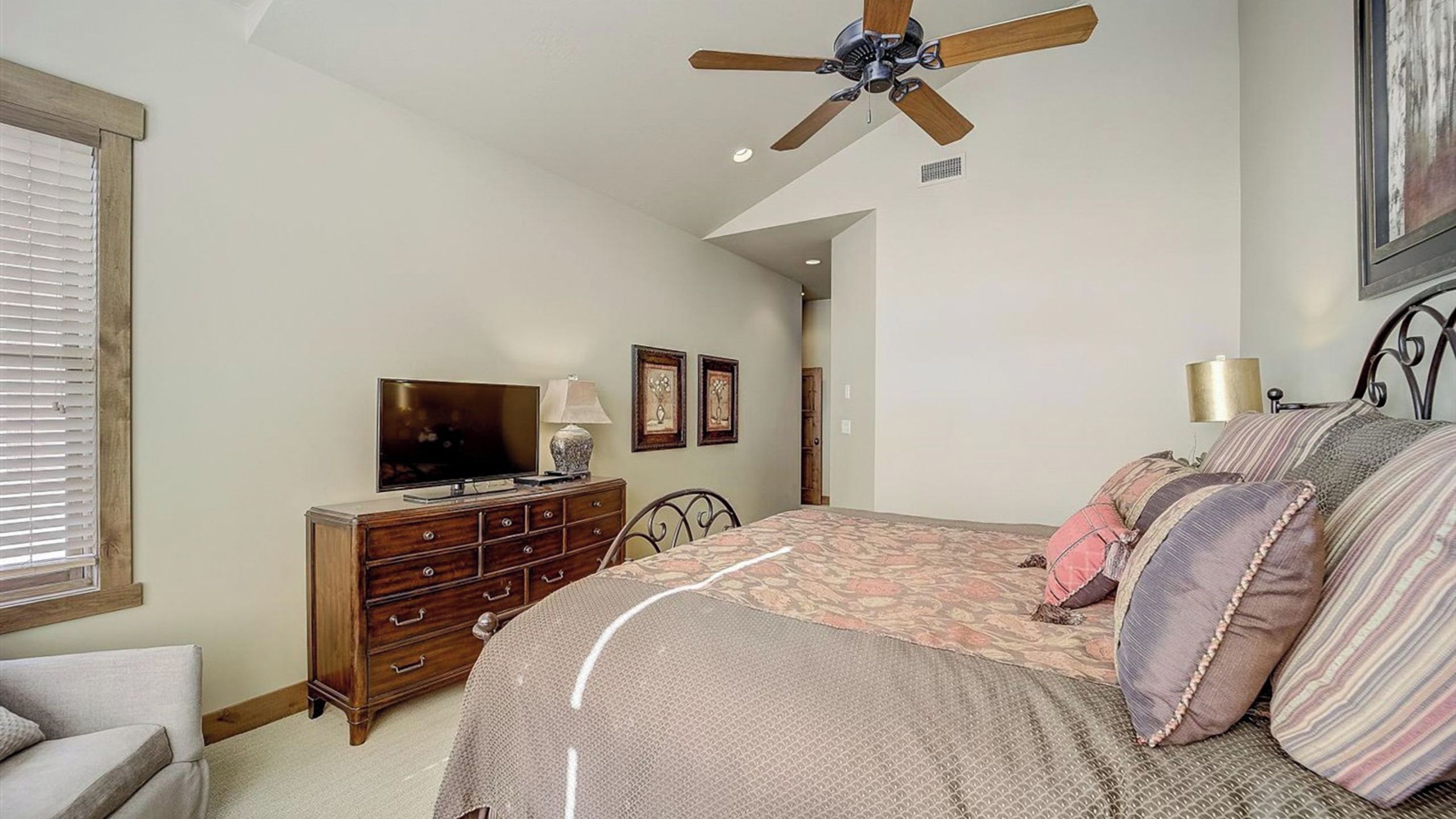 A bedroom with a bed, a dresser, a television, a ceiling fan, and a door leading to another room.