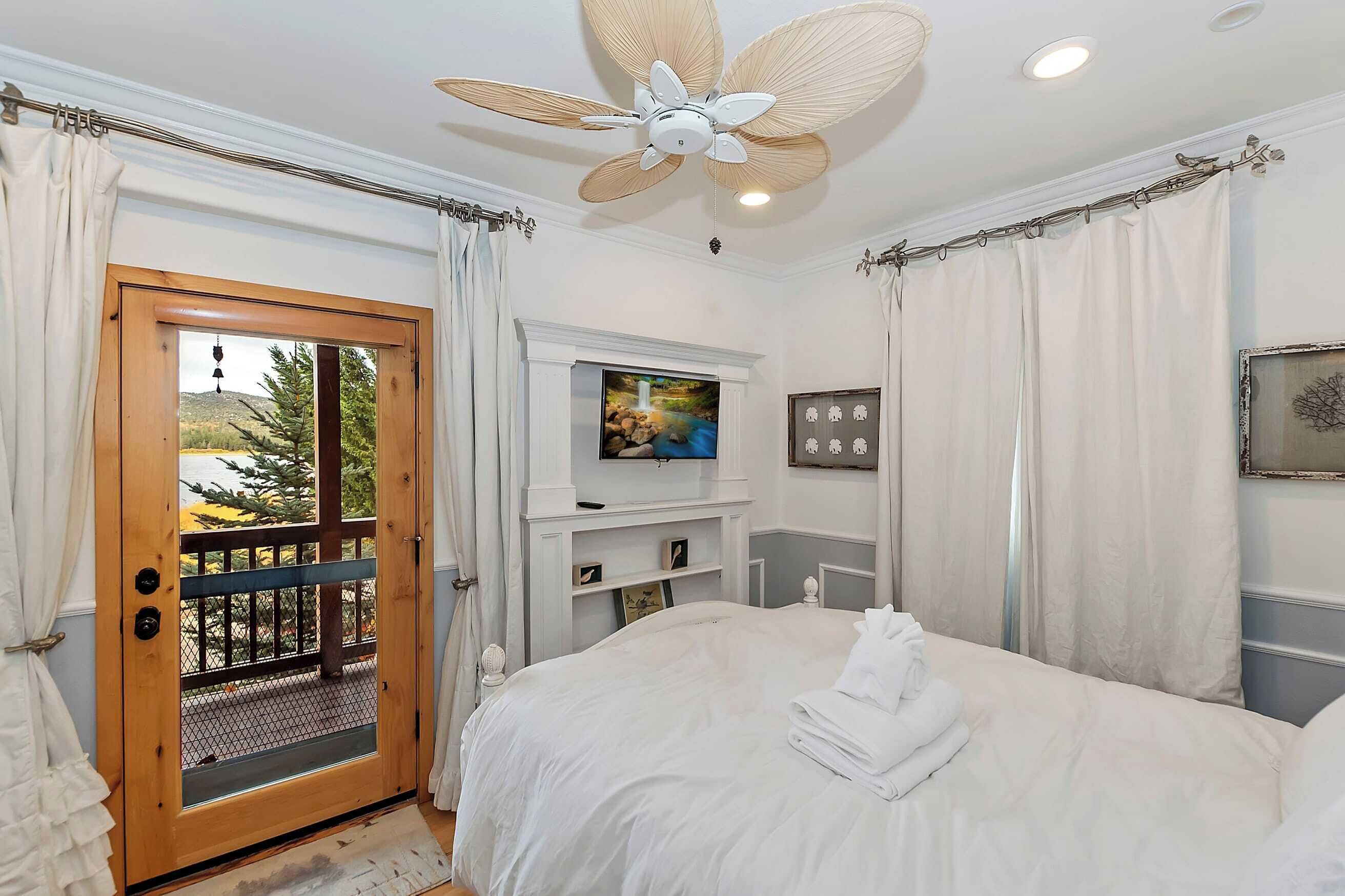A bedroom with a bed, a ceiling fan, a TV, and a view of the outdoors through a door.