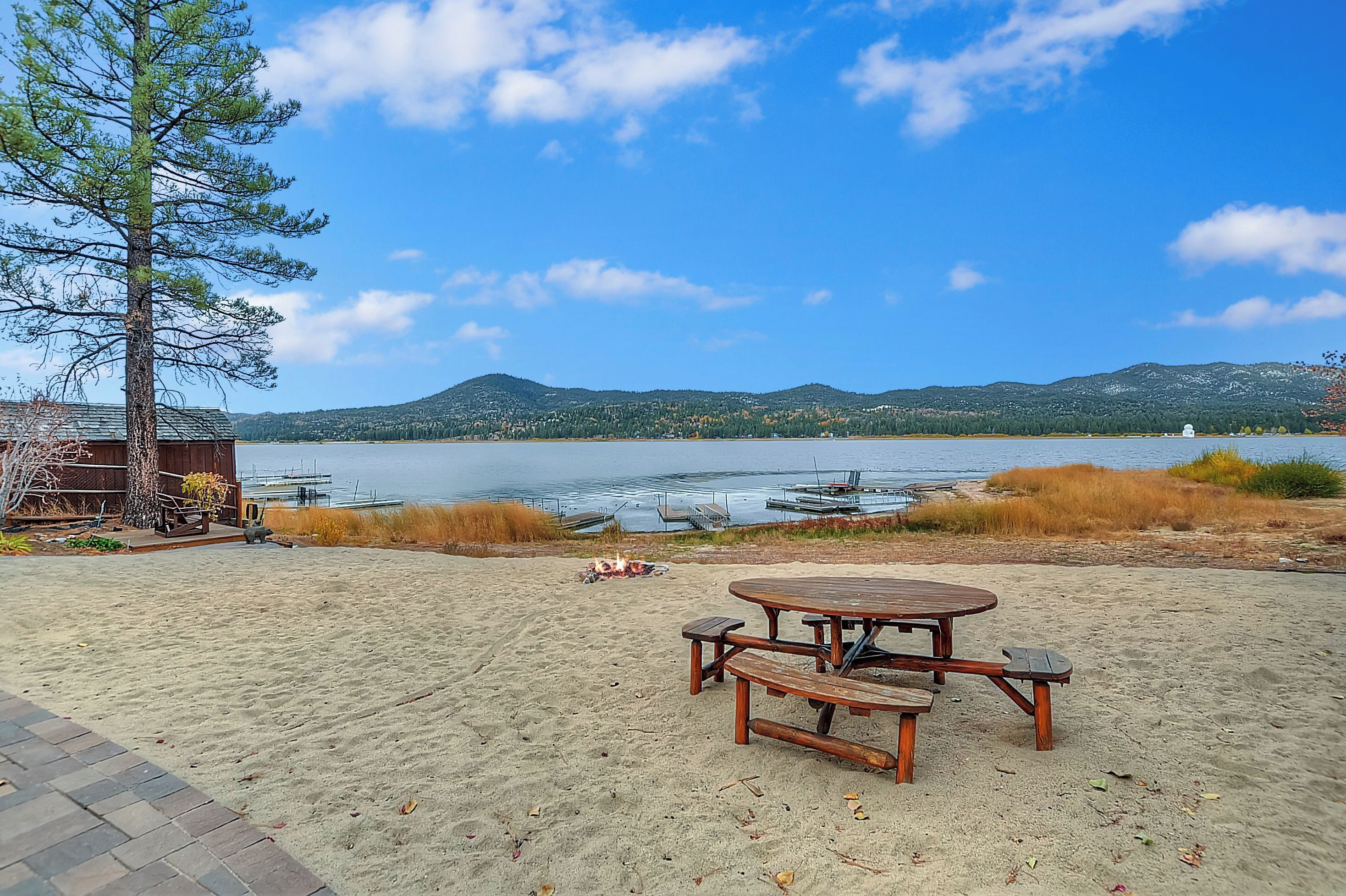 A beach with a wooden picnic table and benches, a fire pit, and a view of the lake and mountains.