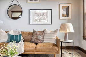 A brown leather sofa with various cushions, a round mirror, a framed abstract artwork, a lamp on a small table, and a window with a view.