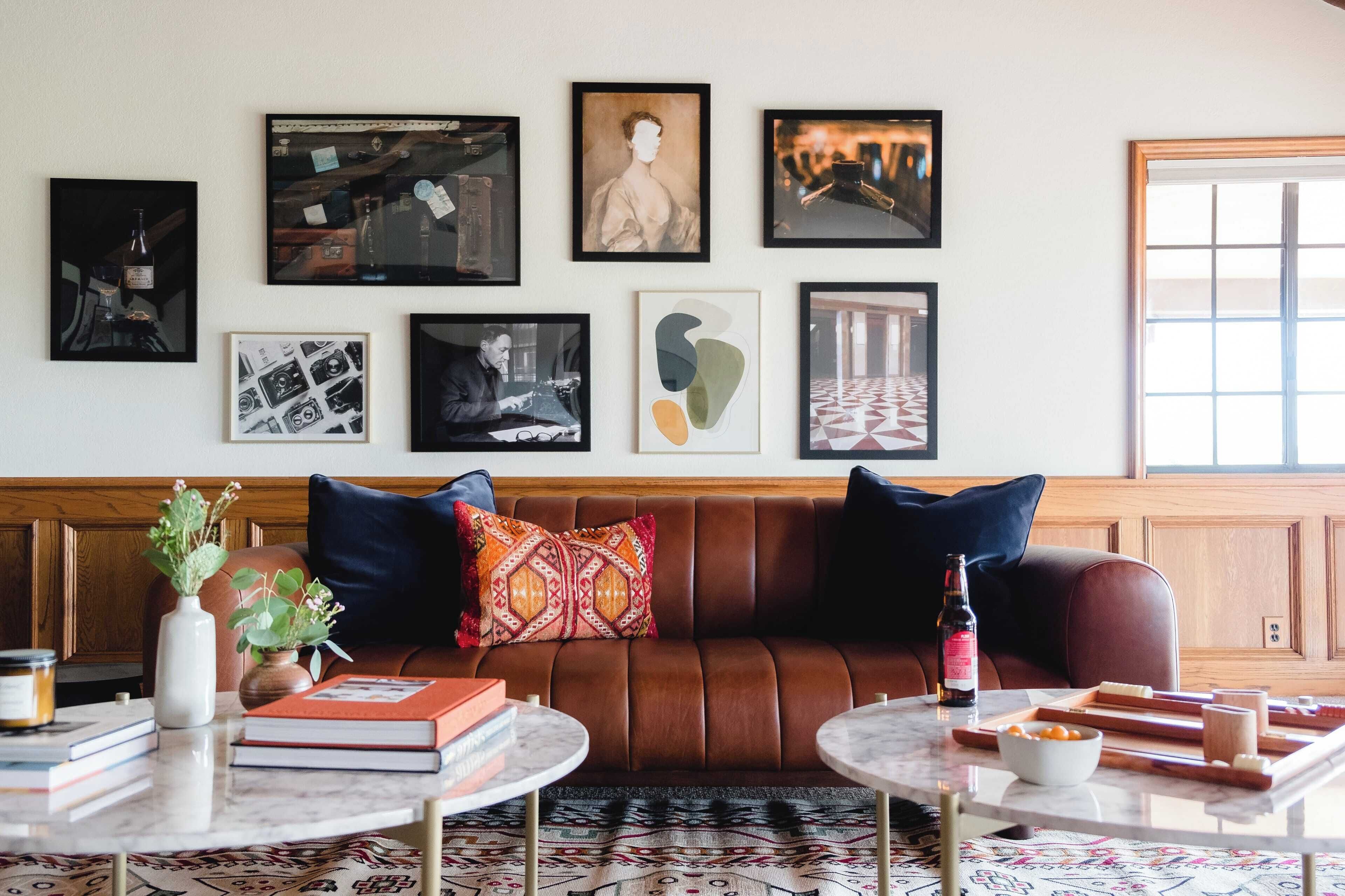 A living room with a brown leather sofa, two round tables, and various framed artworks on the wall.