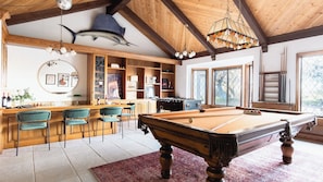 A pool table with a wooden finish, a bar area with stools, and a ceiling with wooden beams and a hanging light fixture.
