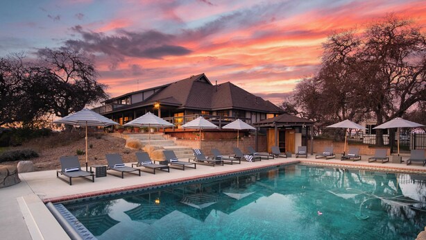 A swimming pool with lounge chairs and umbrellas, surrounded by a building with a sloped roof and a view of a sunset. - Cathedral by Avantstay Charming Secluded Estate w/ Sauna, Pool Table & Views of the Rolling Hills (Templeton)